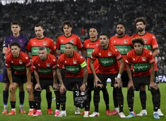 НЕК вдига каре за мача с Левски от Лига Европа NEC Nijmegen football players in red and green kits pose for a group photo on the pitch with a stadium crowd behind them, including a purple-clad goalkeeper on the left.