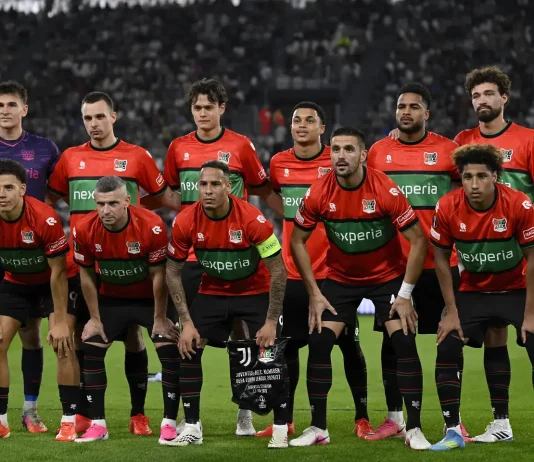 НЕК вдига каре за мача с Левски от Лига Европа NEC Nijmegen football players in red and green kits pose for a group photo on the pitch with a stadium crowd behind them, including a purple-clad goalkeeper on the left.