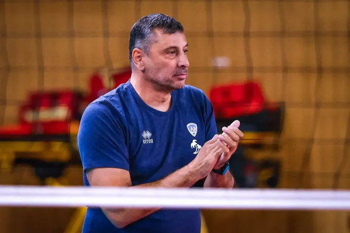 Coach or team staff member in a blue T-shirt clapping in an indoor sports venue, with red seats in the background.
