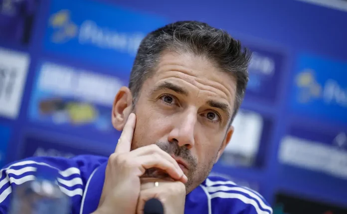Man in a blue tracksuit rests his chin on his hand, appearing thoughtful at a press conference backdrop.