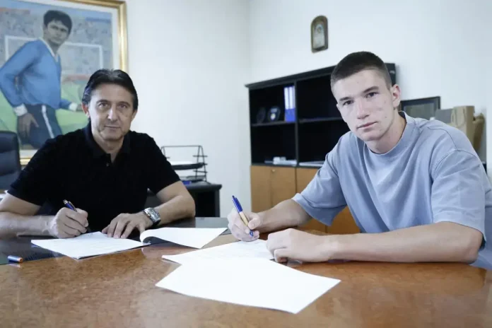 Two men sit at a wooden desk signing papers in an office setting.
