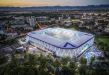 Новият стадион на Левски привлече внимание и в чужбина Aerial view of a modern, illuminated stadium with a white and blue lattice exterior at dusk, surrounded by trees and a cityscape.