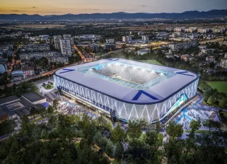 Новият стадион на Левски привлече внимание и в чужбина Aerial view of a modern, illuminated stadium with a white and blue lattice exterior at dusk, surrounded by trees and a cityscape.