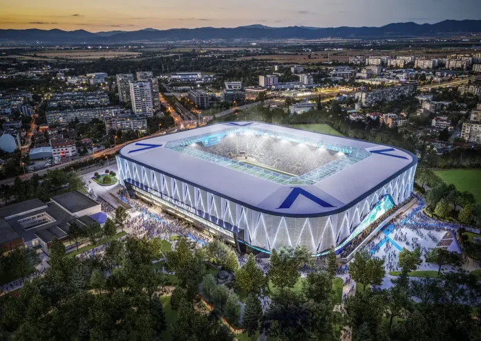 Aerial view of a modern, illuminated stadium with a white and blue lattice exterior at dusk, surrounded by trees and a cityscape.
