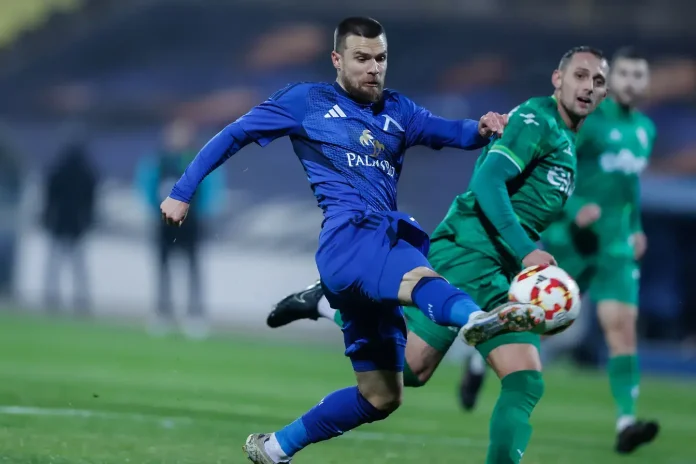 Soccer player in a blue kit kicking a ball during a match, with a green-clad opponent nearby on the field.