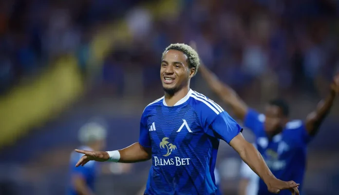 Soccer player in a blue jersey celebrating with arms outstretched on a field; Palms Bet logo on chest.