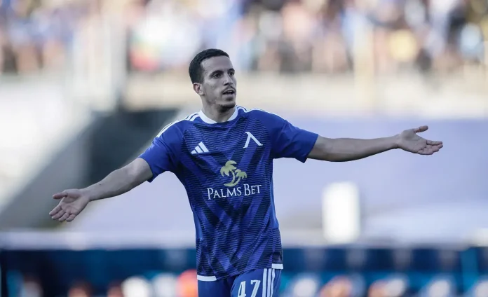 Soccer player in a blue jersey with arms outstretched in celebration on the field, Palms Bet sponsor visible on chest.