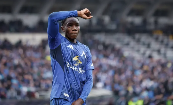 Soccer player in blue kit raising his right arm in celebration on the field with a blurred stadium crowd behind him