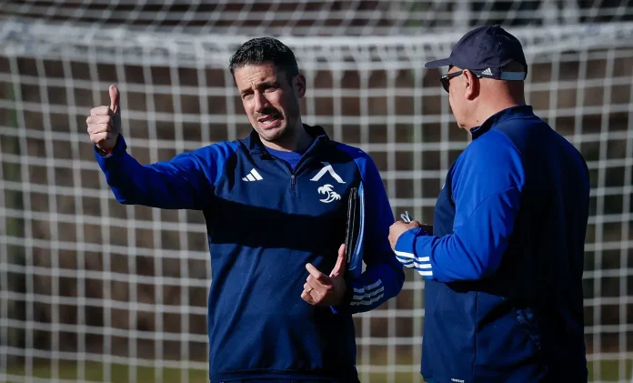 Coach gives a thumbs-up while talking to a teammate on a soccer field, goal net in the background.