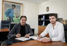 Петко Христов дебютира за Левски, Веласкес направи 10 промени в състава Two men sit at a glossy conference table in an office, signing documents with pens as they look at the camera, one in a dark blazer and the other in a white shirt; a large framed painting of a soccer player hangs on the wall behind them.