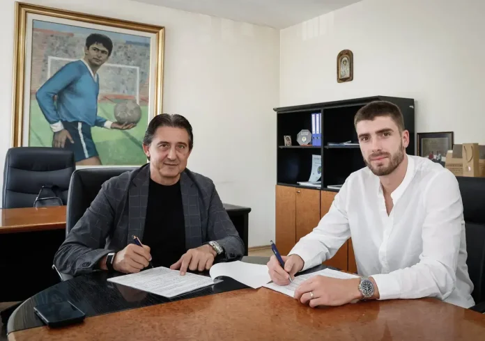 Two men sit at a glossy conference table in an office, signing documents with pens as they look at the camera, one in a dark blazer and the other in a white shirt; a large framed painting of a soccer player hangs on the wall behind them.