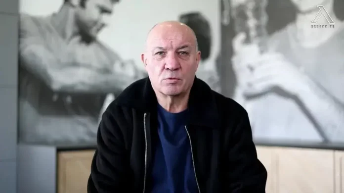 Bald man in a black jacket speaks to the camera in a modern office, with a grayscale mural behind him.