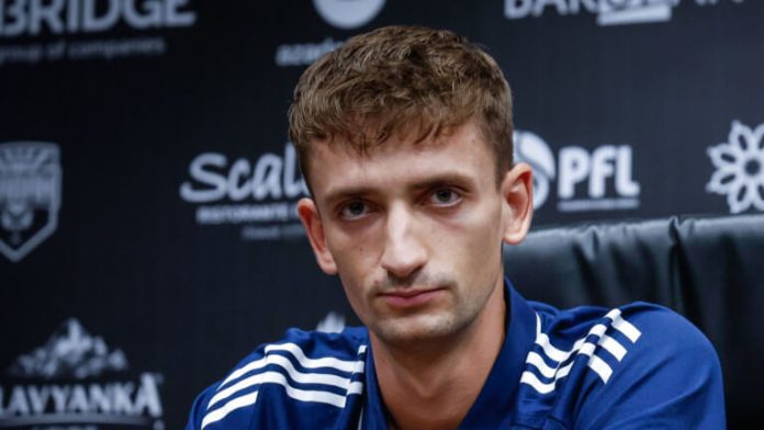 Man in a blue sports jacket sits at a press conference, looking at the camera with a neutral expression, backdrop covered in logos.