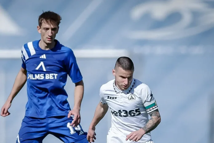 Two soccer players compete for the ball on a sunny field, one in a blue kit and one in white with a captain's armband on his left arm.
