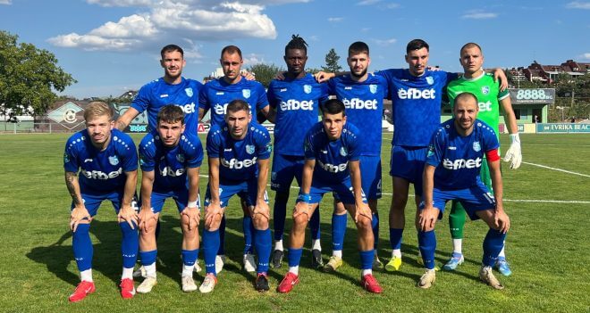 Eleven male football players in blue kits pose for a team photo on a sunny grass field, with a green-clad goalkeeper on the right.