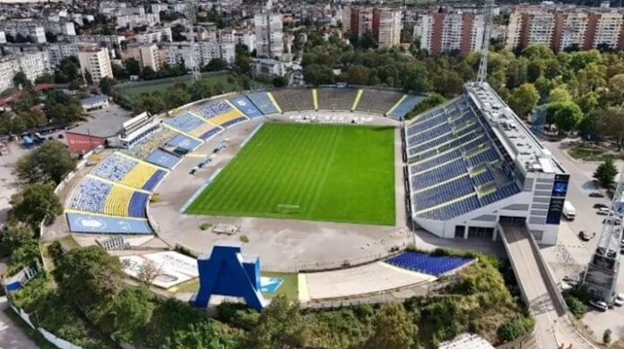 Aerial view of a stadium with a bright green pitch and blue-yellow seating surrounding a circular track-like layout, in a city setting.