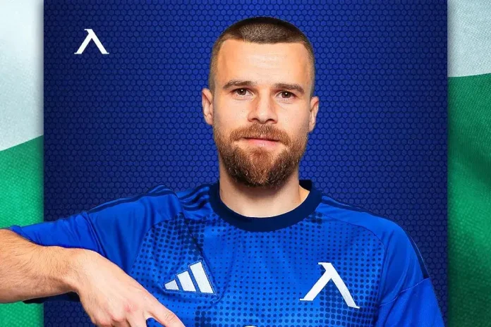 Portrait of a man with short hair and beard wearing a blue athletic jersey with white logos against a blue backdrop.