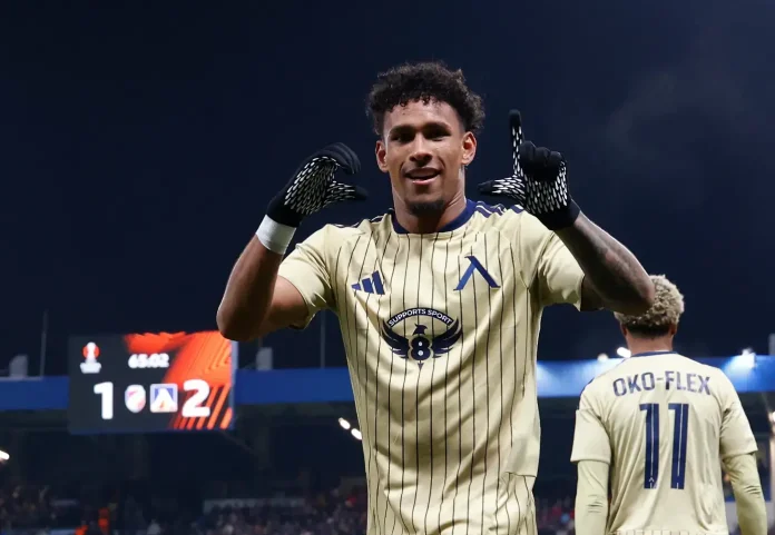 Footballer in a beige pinstripe kit celebrates with hands raised, number 11 OKO-FLEX on the back, stadium crowd behind him.