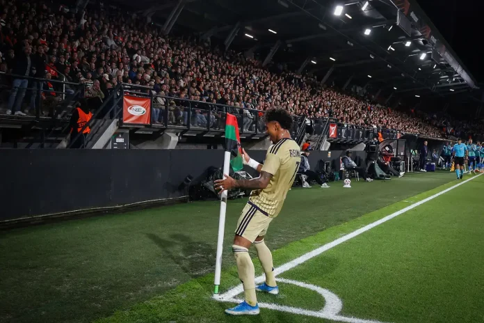Soccer player in a beige kit leans on the corner flag near the sideline as a packed stadium watches.