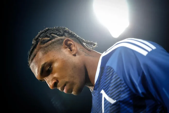 Profile view of a male athlete with cornrow braids in a blue jersey, illuminated by bright stadium lights.