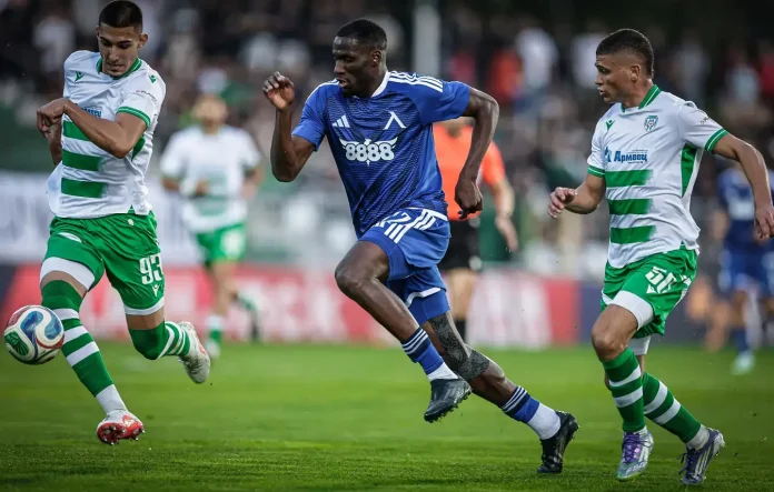 Blue-team player in mid-run dribbles the ball while two green-and-white striped players pursue him on a soccer field.