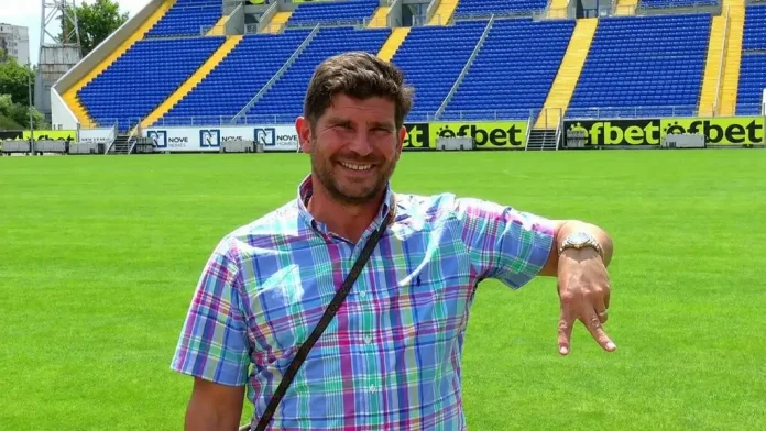 Smiling man in a pastel plaid shirt stands on a green soccer field with blue and yellow stadium seats in the background, pointing downward with his right hand.