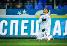 Шейхове изкушават Евертон Бала Soccer player in a white kit with number 17 kneels on the grass, hand over chest, looking up in celebration or prayer on a pitch with blue advertising boards behind.