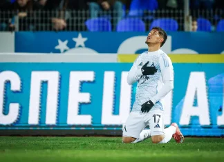 Шейхове изкушават Евертон Бала Soccer player in a white kit with number 17 kneels on the grass, hand over chest, looking up in celebration or prayer on a pitch with blue advertising boards behind.