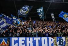 Скандал: НЕК скочи на Левски и сезира УЕФА! Large stadium crowd cheering with raised arms, waving blue flags and banners behind a barrier.