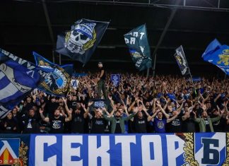 Скандал: НЕК скочи на Левски и сезира УЕФА! Large stadium crowd cheering with raised arms, waving blue flags and banners behind a barrier.