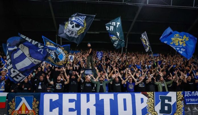 Large stadium crowd cheering with raised arms, waving blue flags and banners behind a barrier.