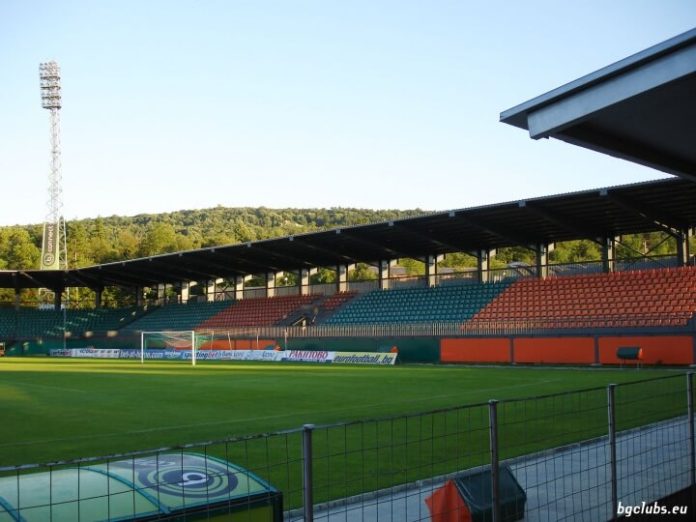 Empty football stadium with a green pitch, goal, advertising boards, and multicolored seating under a clear blue sky; fence in the foreground.