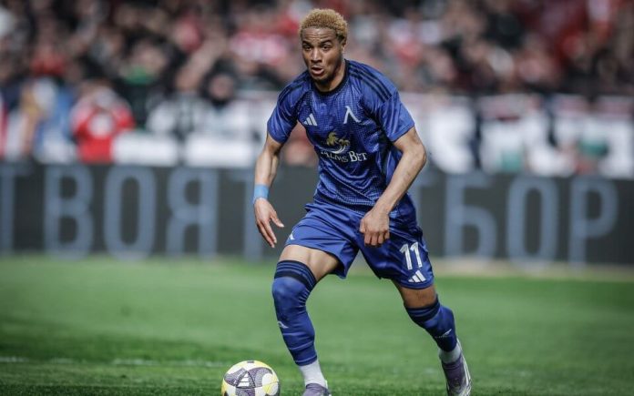 Soccer player in a blue kit dribbles the ball on a grass pitch during a match, with fans in the stands.