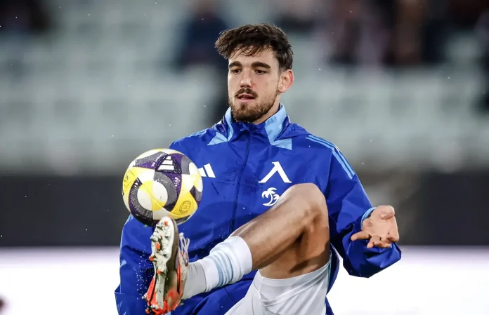 Soccer player in a blue training jacket traps a yellow-purple ball with his foot mid-action on the field.