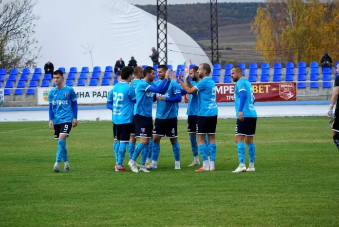 Soccer players in blue kits form a huddle on a green field before a match with empty blue seats behind them.