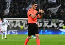 Съдията за гостуването на Левски на НЕК – кошмар за Лудогорец Soccer referee in an orange shirt and black shorts on the field, giving a thumbs-up gesture as players and a crowd look on in the background.