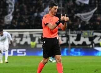 Съдията за гостуването на Левски на НЕК – кошмар за Лудогорец Soccer referee in an orange shirt and black shorts on the field, giving a thumbs-up gesture as players and a crowd look on in the background.