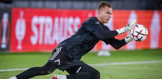 Светослав Вуцов Левски оглави престижна класация Soccer goalkeeper in a black kit diving to his left, reaching for a ball near the goal line on a green pitch with red advertising boards in the background.