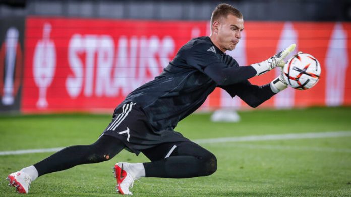 Soccer goalkeeper in a black kit diving to his left, reaching for a ball near the goal line on a green pitch with red advertising boards in the background.