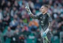 Светослав Вуцов е номер едно в Лига Европа Soccer goalkeeper in a gray kit raises his right index finger while standing on the pitch, with a blurred crowd in the stands behind him.