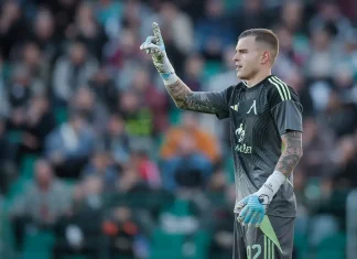 Светослав Вуцов е номер едно в Лига Европа Soccer goalkeeper in a gray kit raises his right index finger while standing on the pitch, with a blurred crowd in the stands behind him.