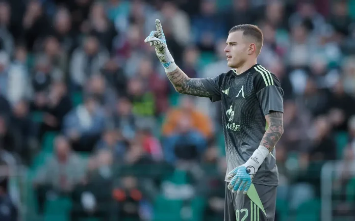 Soccer goalkeeper in a gray kit raises his right index finger while standing on the pitch, with a blurred crowd in the stands behind him.