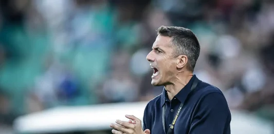 Треньорът на Левски има история с НЕК Coach in a dark navy polo shouts and claps on the sidelines at a sports stadium, wearing a lanyard and watch.