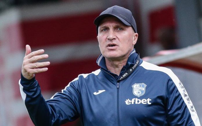 Coach in a navy blue jacket with a team crest speaks and gestures on the sideline while wearing a cap.