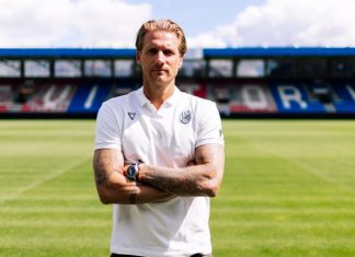 Треньорът на Виктория Пилзен: Знаем слабите места на Левски Male soccer coach standing on a grassy pitch with folded arms, wearing a white polo shirt and a watch, empty stadium seating in the background.