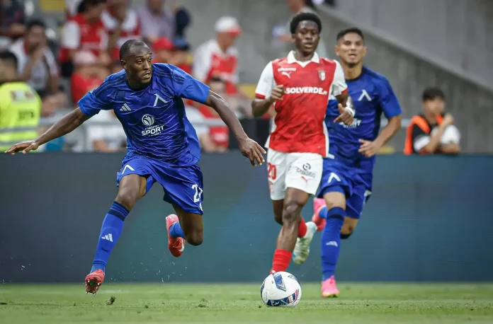 Soccer player in blue kit lunges toward the ball as other players in blue and red kits chase behind on a grassy field.