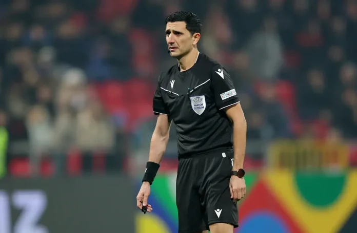 Male football referee in black kit standing on the pitch during a match, with a UEFA badge on his chest.