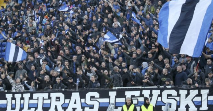 Packed stadium crowd of football fans in blue and white, waving flags and raising arms behind a bold banner.