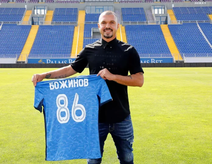 Man in a black polo shirt stands on a soccer field, holding up a blue jersey with the number 86 and Cyrillic text, with blue and yellow stadium seats in the background.
