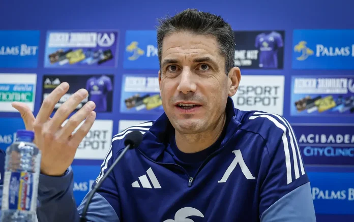 Man in a blue athletic jacket speaks at a press conference with a microphone in front; sponsor boards are blurred in the background.